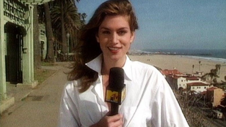 Cindy Crawford, wearing a white collared blouse with her hair down and blowing in the wind, holds the MTV News microphone while reporting near houses and palm trees next to the beach on "House of Style."
