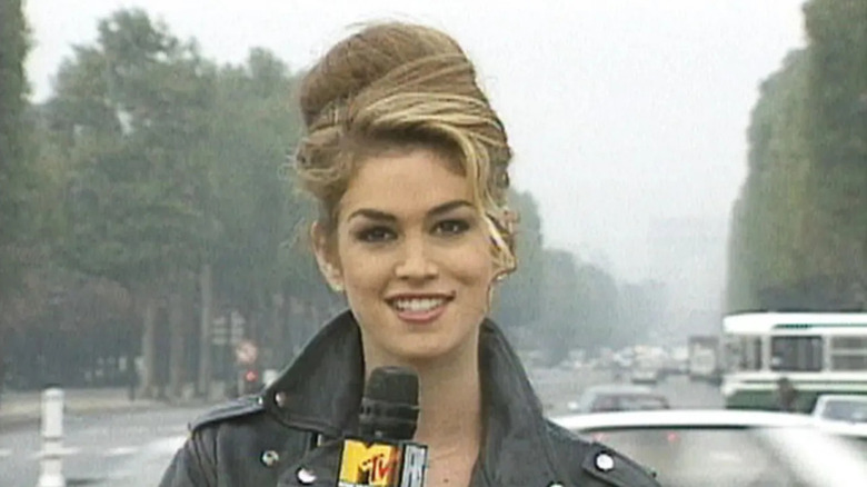 Cindy Crawford, wearing a black leather jacket with her hair in a beehive up-do, holds the MTV News microphone while street reporting on "House of Style."
