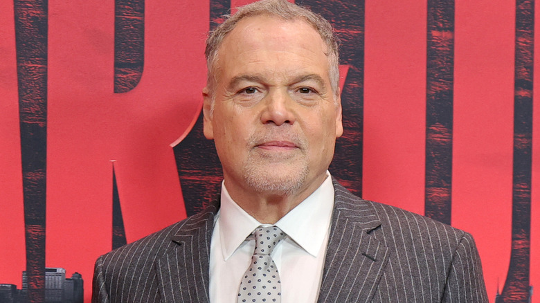 Vincent D'Onofrio with gray hair and a goatee wearing a gray pinstriped suit and a gray polka-dot tie and a white shirt.