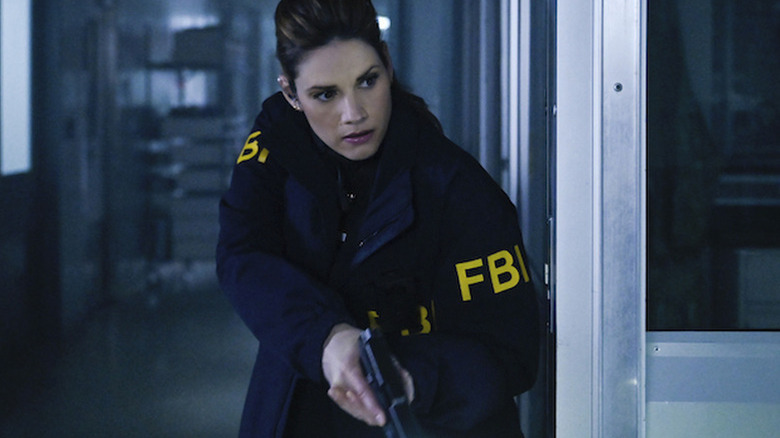 Missy Peregrym as special agent Maggie Bell, wearing her blue uniform FBI jacket with "FBI" in yellow lettering on the sleeves, holding her gun low and looking around a corner in FBI
