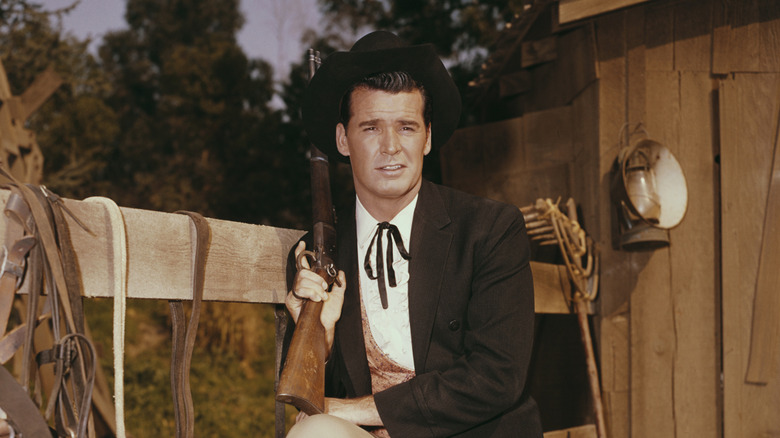 Bret Maverick posing with his shotgun while sitting on a bale of hay next to a wooden fence