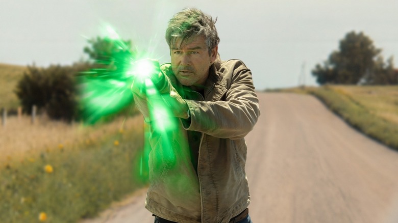 Kyle Chandler as Hal Jordan with graying hair and stubble wearing a brown leather jacket holding his arm out, there is a bright green light glowing from a ring on his finger.