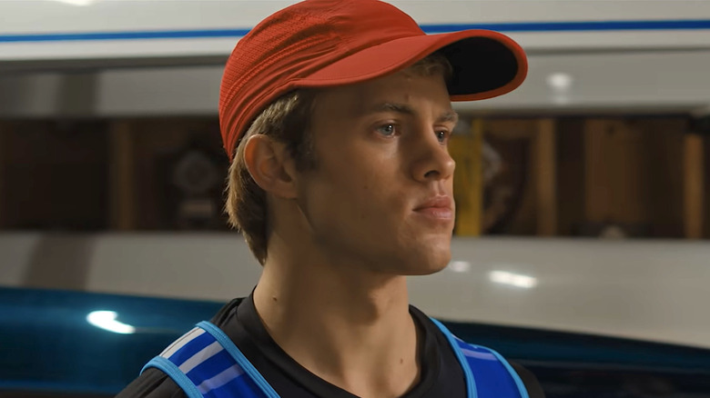 Josh Regis wearing a red hat, black shirt, and blue best looking to his left in Crew Girl