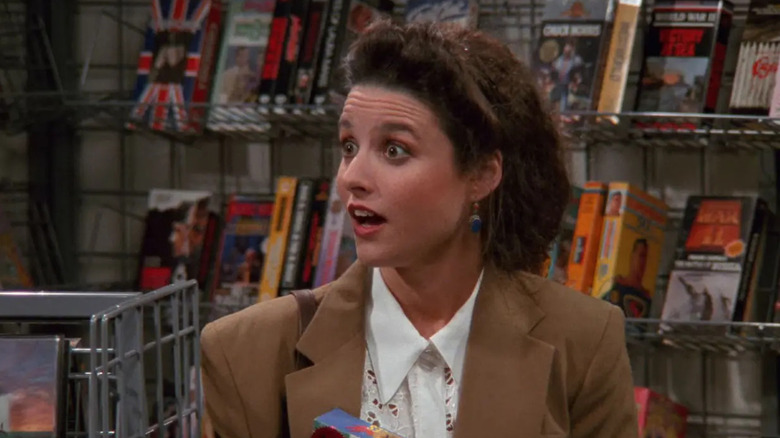 Elaine Benes (Julia Louis-Dreyfus) wears a tan blazer over a white blouse, holding a box and looking surprised inside a video rental store on Seinfeld