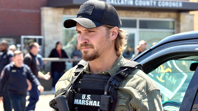 Kayce Dutton in a black U.S. Marshal vest and a black hat holding a rifle in front of a car in Marshals