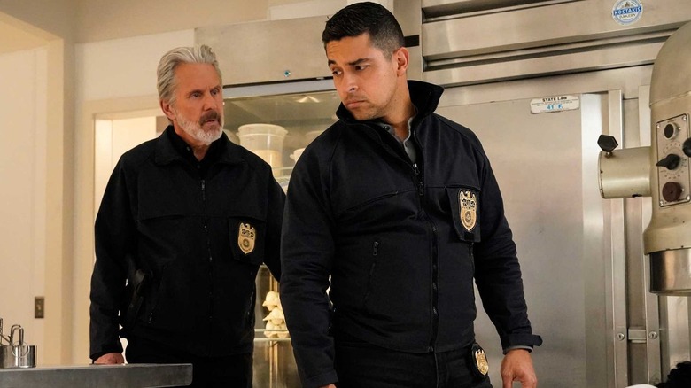 Özel ajan Alden Parker rolünde Gary Cole, özel ajan Nicholas Torres rolünde Wilmer Valderrama'nın arkasında yürüyor; omzunun üzerinden Parker'a bakıyor; ikisi de endüstriyel bir mutfakta, sol göğüs cebinde rozetli NCIS siyah kazakları giyiyor. 
