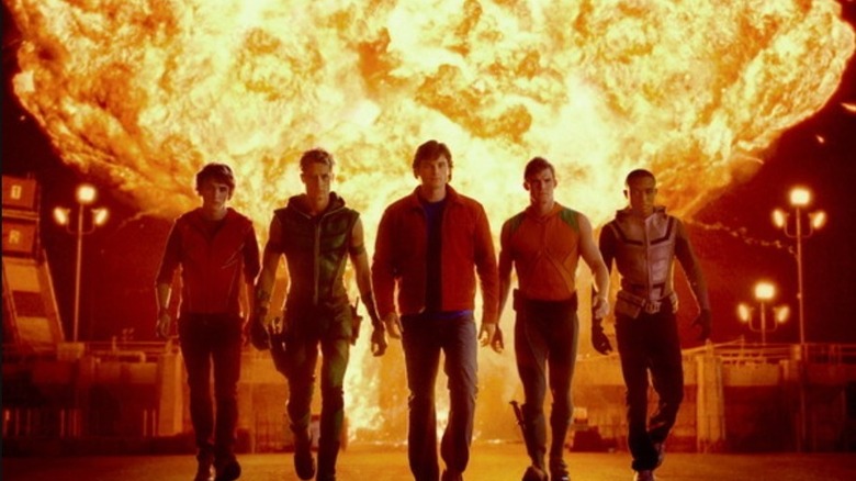 Five young superheroes—Flash (Kyle Galner), Green Arrow (Justin Hartley), Clark Kent (Tom Welling), Aquaman (Alan Ritchson), and Cyborg (Lee Thompson Young)—walk forward side-by-side as a massive explosion erupts directly behind them in the series "Smallville."