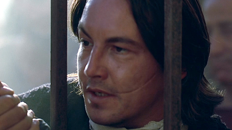 A man with long brown hair stands behind bars. There is a large scar curved across his left cheek.
