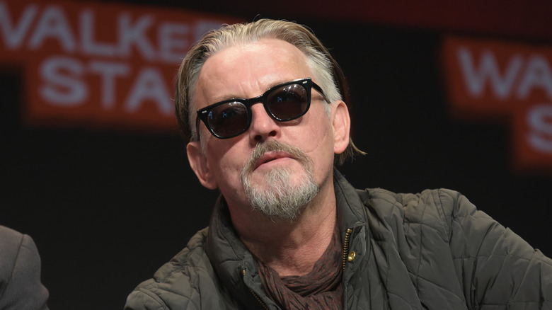 An older man with swept-back graying hair and a goatee wearing ray ban sunglasses and a quilted jacket. There is a display behind him with red outlined white text that reads "Walker Stalker"