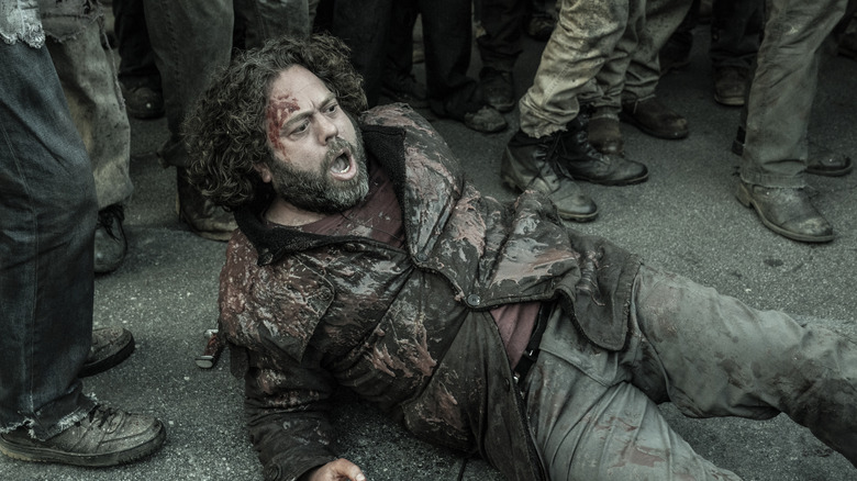 A man with large curly brown hair and a graying beard is lying on the floor with a small amount of blood on his face and a blood stained jacket with a red t-shirt underneath. The angle of the image shows him surrounded by a crowd of people in ruined trousers and shoes, but it doesn't show their upper bodies.