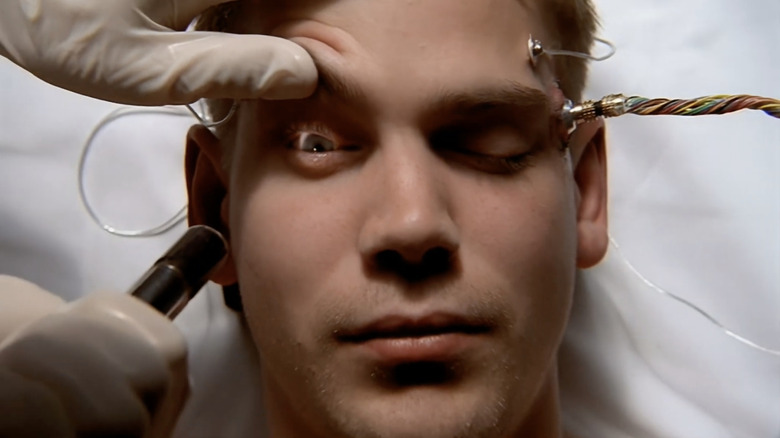 Scott Bairstow as Thomas Hobbes in "Harsh Realm" lying in a hospital bed as someone wearing white gloves opens his eyelid. There is a large cable plugged into his forehead.