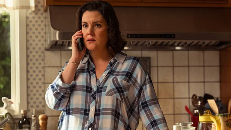 Shauna (Melanie Lynskey) in a checkered shirt stands in a kitchen, holding a phone to her ear, looking concerned in Yellowjackets