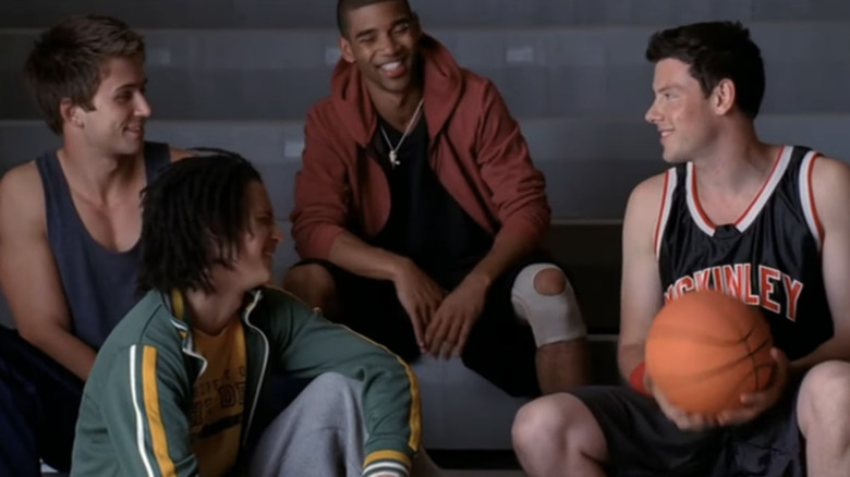 Brandon Sklenar as a high schooler sitting on the bleachers with two background actors also playing high schoolers and Cory Monteith as Finn in a McKinley High School Basketball uniform in Glee
