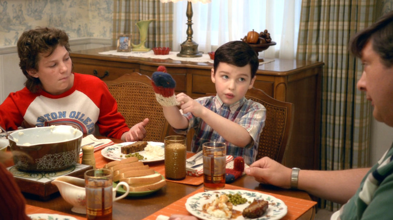 Georgie and George Sr. looking at young Sheldon (who's putting on a mitten) at the dinner table, in the Young Sheldon pilot