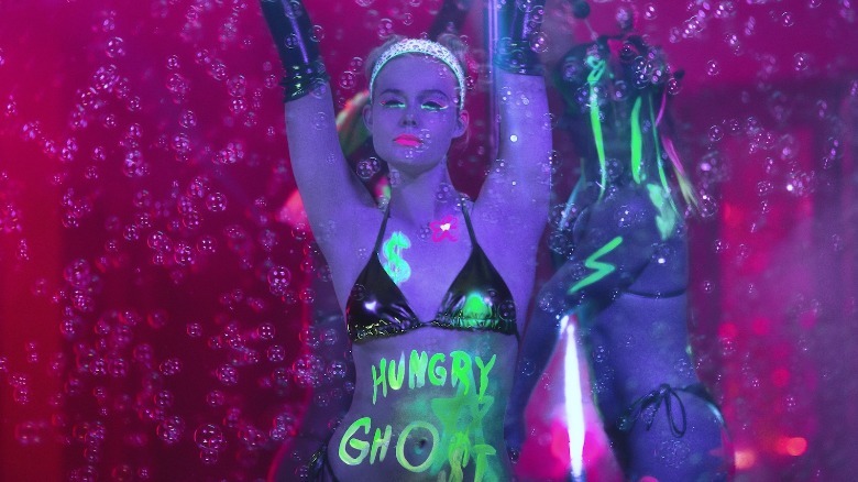 Elle Fanning como Margo em Margo's Got Money Trouble usa um biquíni e tem HUNGRY GHOST escrito em seu torso com tinta glitter.